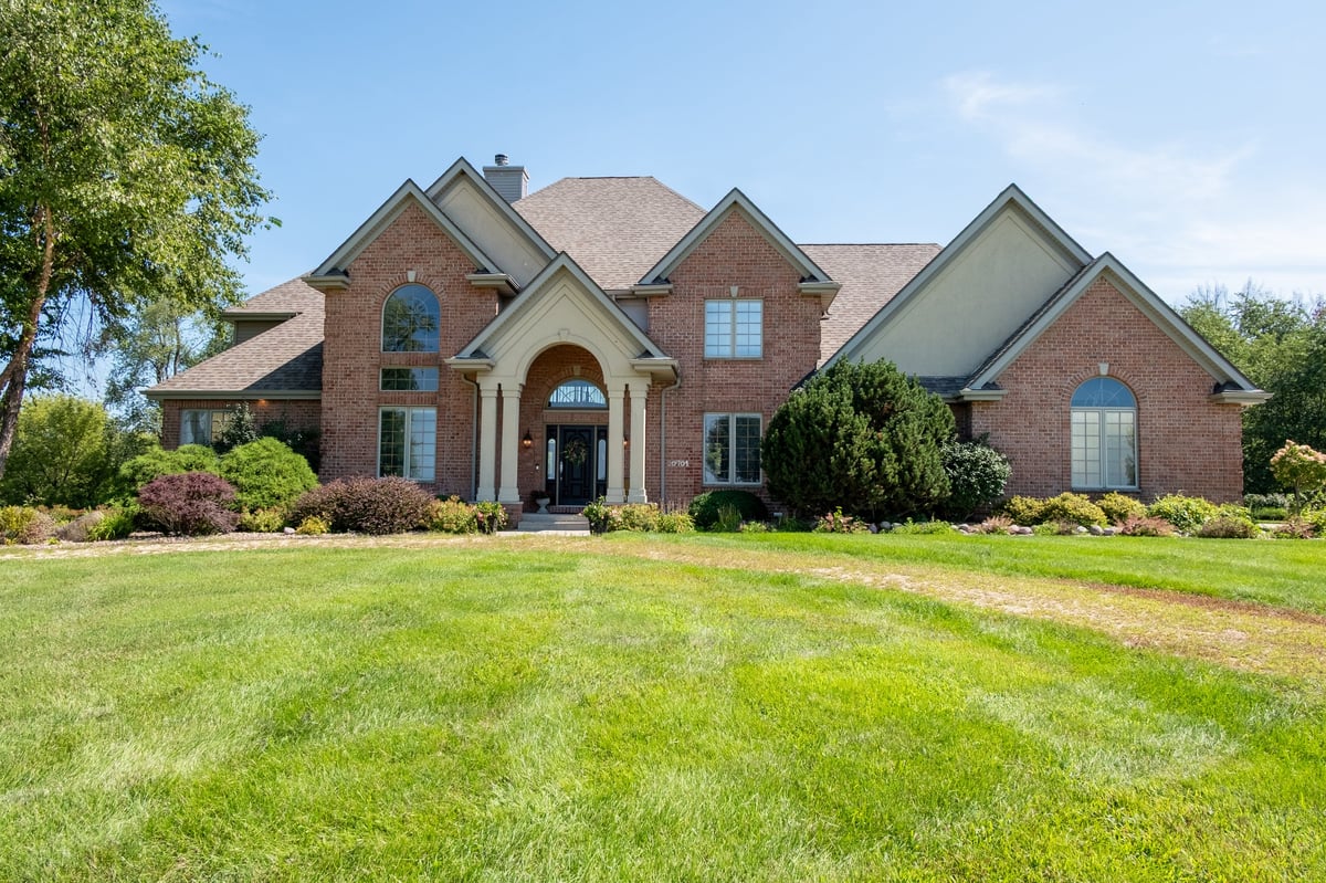 Large two-story brick house with arched entryway, multiple gables, and manicured lawn surrounded by green landscaping.