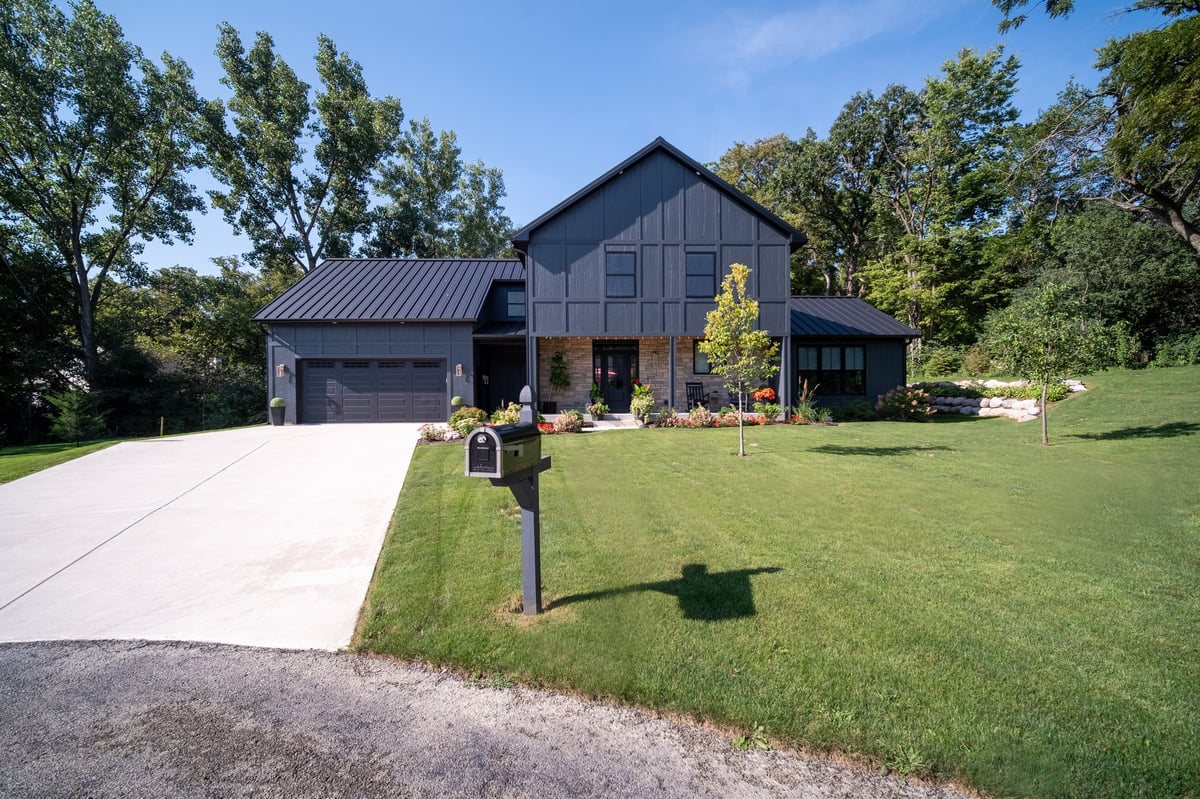 Modern black barn-style house with metal roof, two-car garage, mailbox, and manicured lawn surrounded by trees
