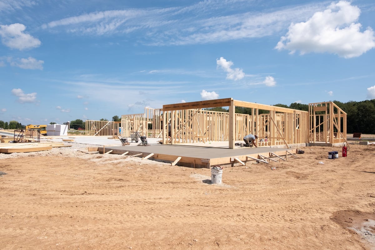 New home under construction with wooden framing on a foundation in a dirt lot under clear blue sky.